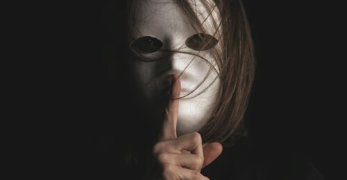 A person wearing a blank white masking, holding a finger in front of their mouth to indicate silence.